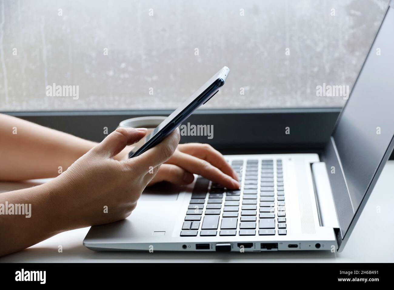 Femme avec un smartphone assis au clavier de l'ordinateur portable près de la fenêtre. Concept de communication en ligne, travail au bureau ou à domicile et paiement Banque D'Images