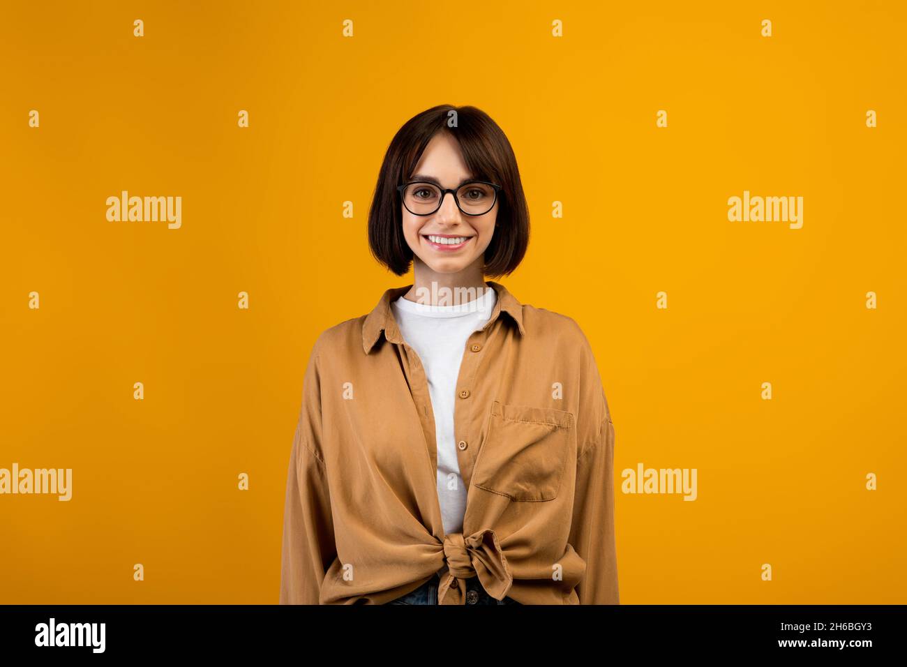Une étudiante gaie portant des lunettes et souriant à l'appareil photo, posant sur un fond jaune de studio.Portrait de la bonne dame avec bob coupe de cheveux Banque D'Images