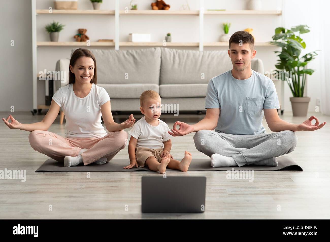 Yoga en ligne.Bonne famille jeune avec bébé enfant méditant ensemble à la maison avec ordinateur portable, parents joyeux assis dans la position Lotus, regarder la vidéo Banque D'Images