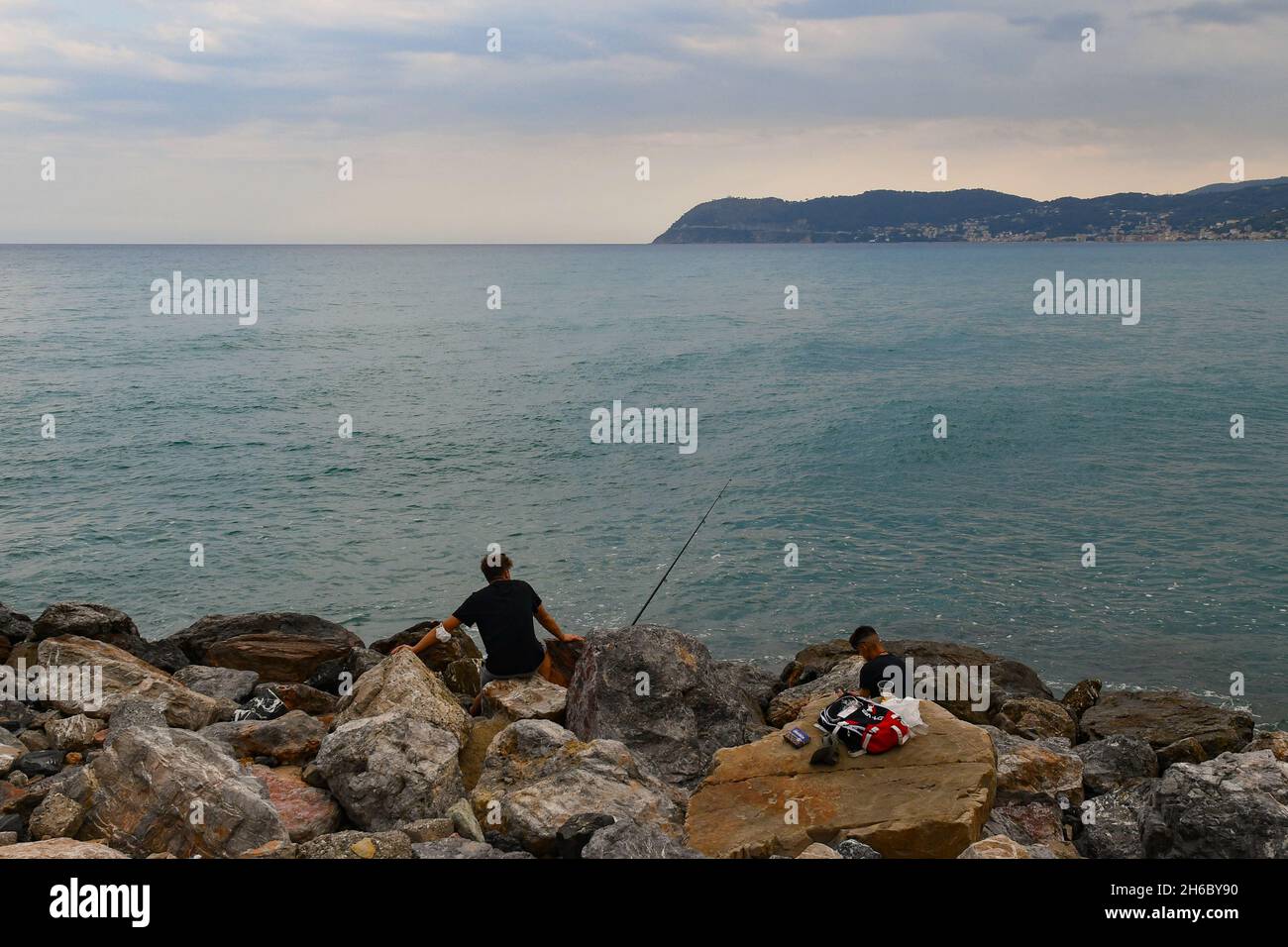 Deux garçons pêchant à partir de la mer rocheuse avec le promontoire de Capo Mele au-dessus de l'horizon de la mer par une journée nuageux, Alassio, Savona, Ligurie, Italie Banque D'Images