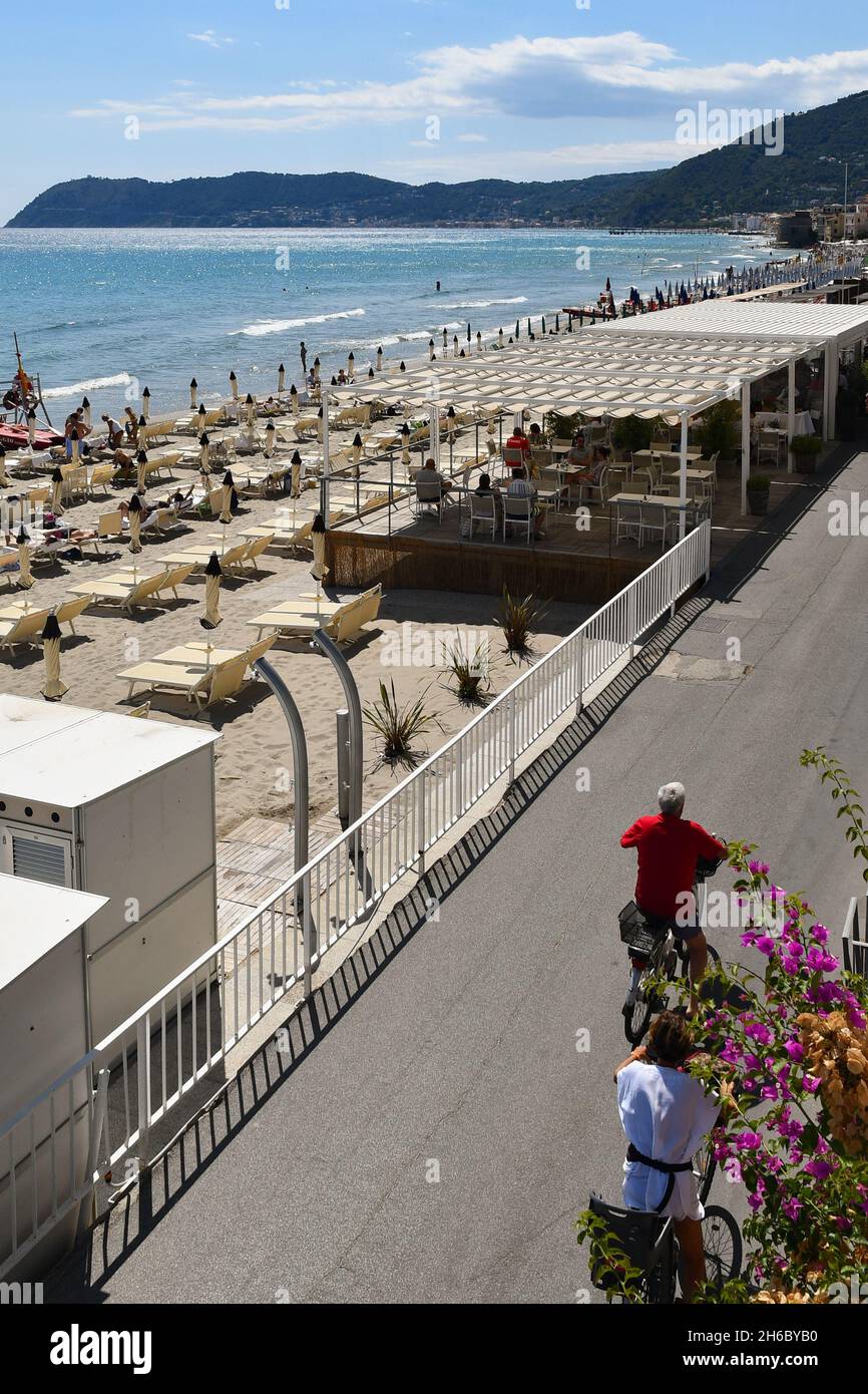 Vue imprenable sur la promenade de la mer avec restaurant de plage sous pergolas, touristes en vélo et Cap Capo Mele en arrière-plan, Alassio, Savona Banque D'Images