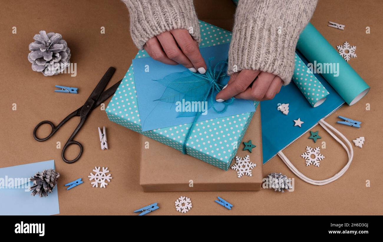 Gros plan les mains de femme emballant et emballant les boîtes-cadeaux de Noël avec du papier bleu, ajoutant enveloppe et feuilles de squelette, vue en grand angle Banque D'Images