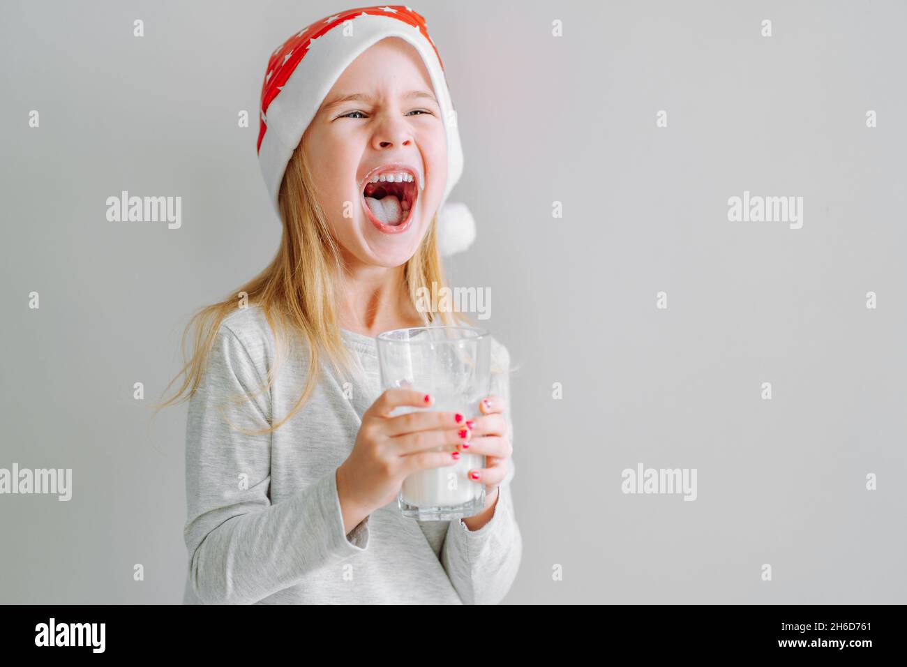 Portrait d'une petite fille en pyjama et chapeau de père noël riant avec verre de lait sur fond clair. Banque D'Images