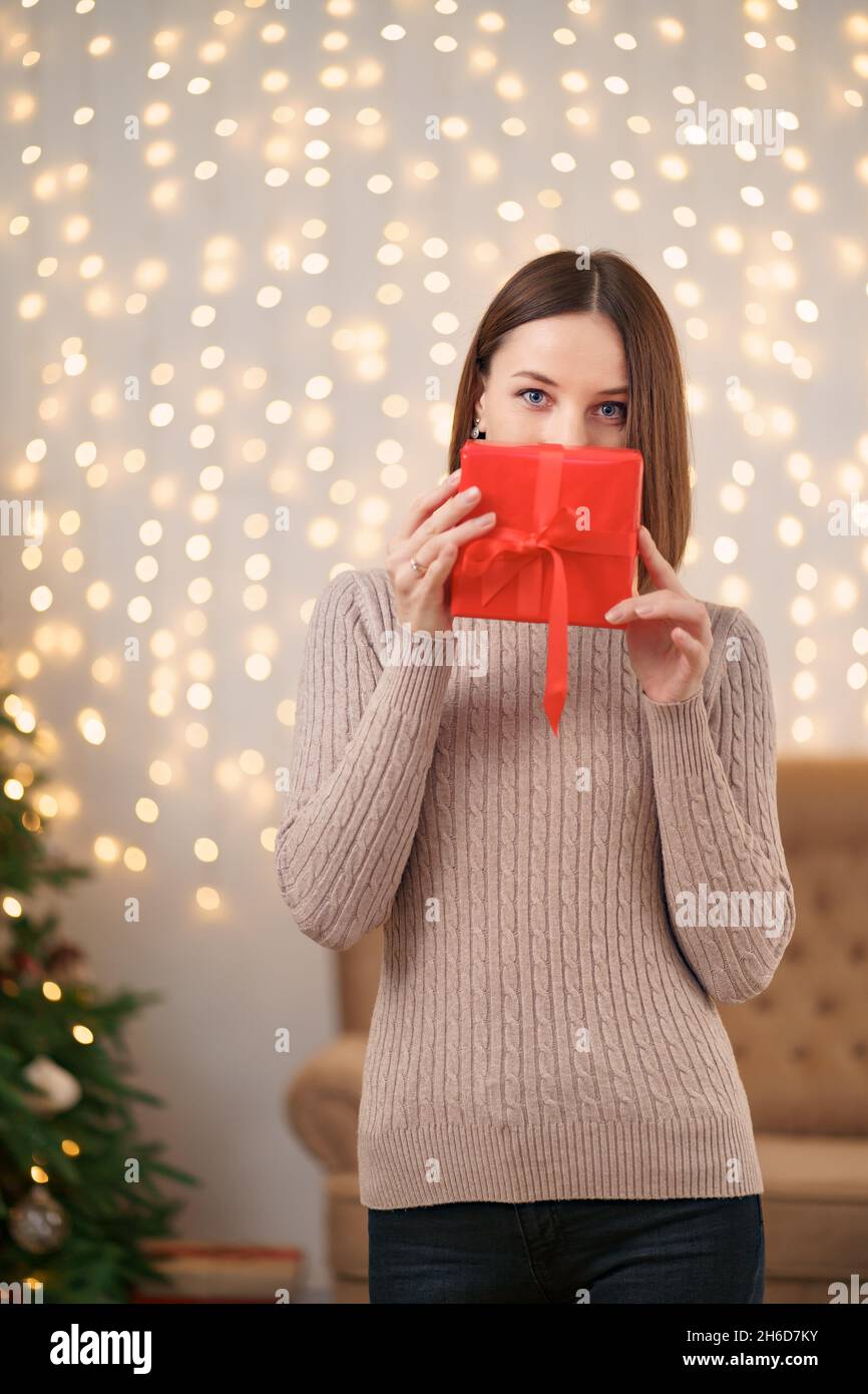 Portrait de la jeune femme heureuse lèvres rouges se cachant derrière la boîte cadeau emballée.Gros plan la femme satisfaite a reçu la boîte actuelle.Arrière-plan des lumières de Noël festives. Banque D'Images