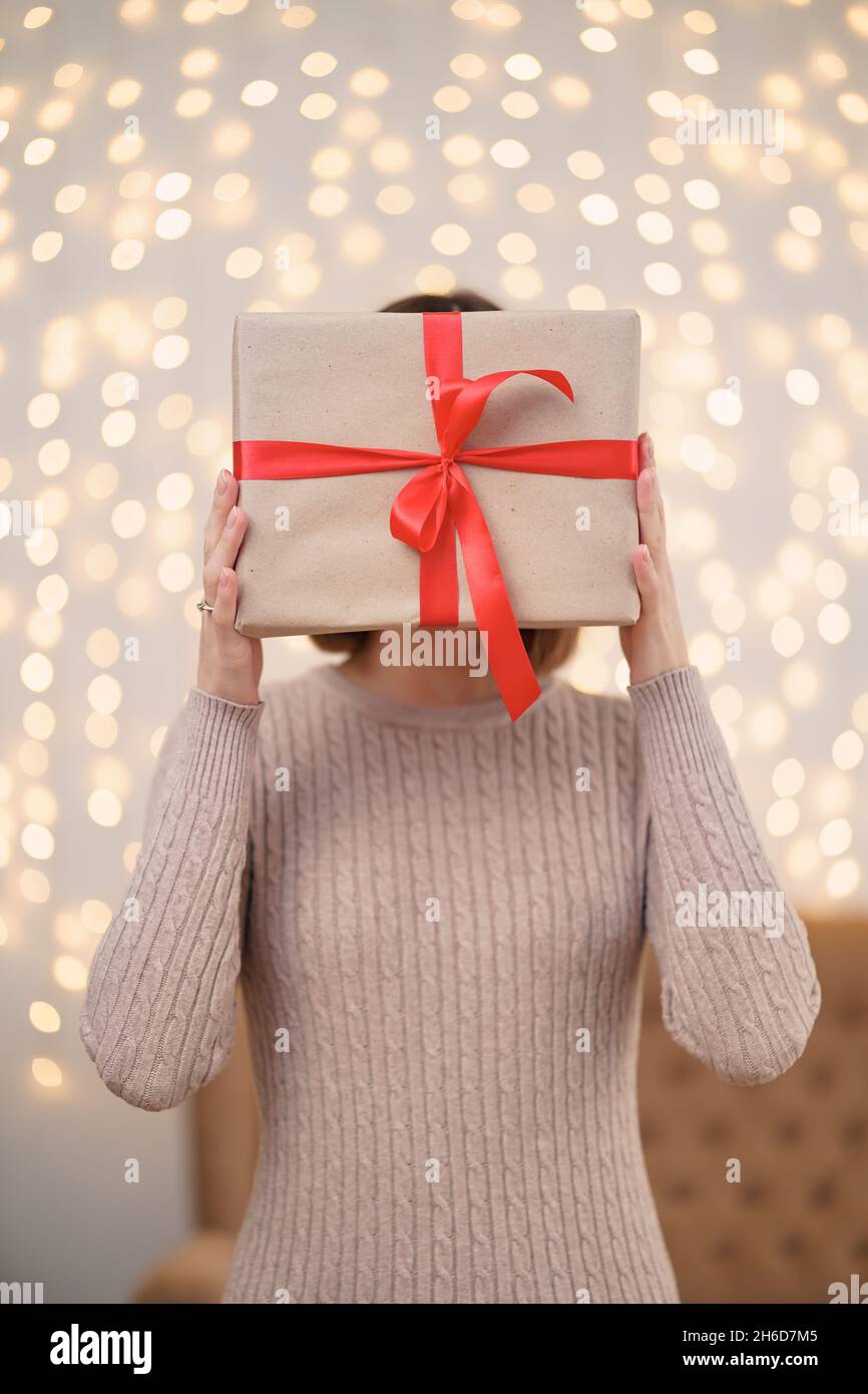 Portrait de la jeune femme heureuse lèvres rouges se cachant derrière la boîte cadeau emballée.Gros plan la femme satisfaite a reçu la boîte actuelle.Arrière-plan des lumières de Noël festives. Banque D'Images