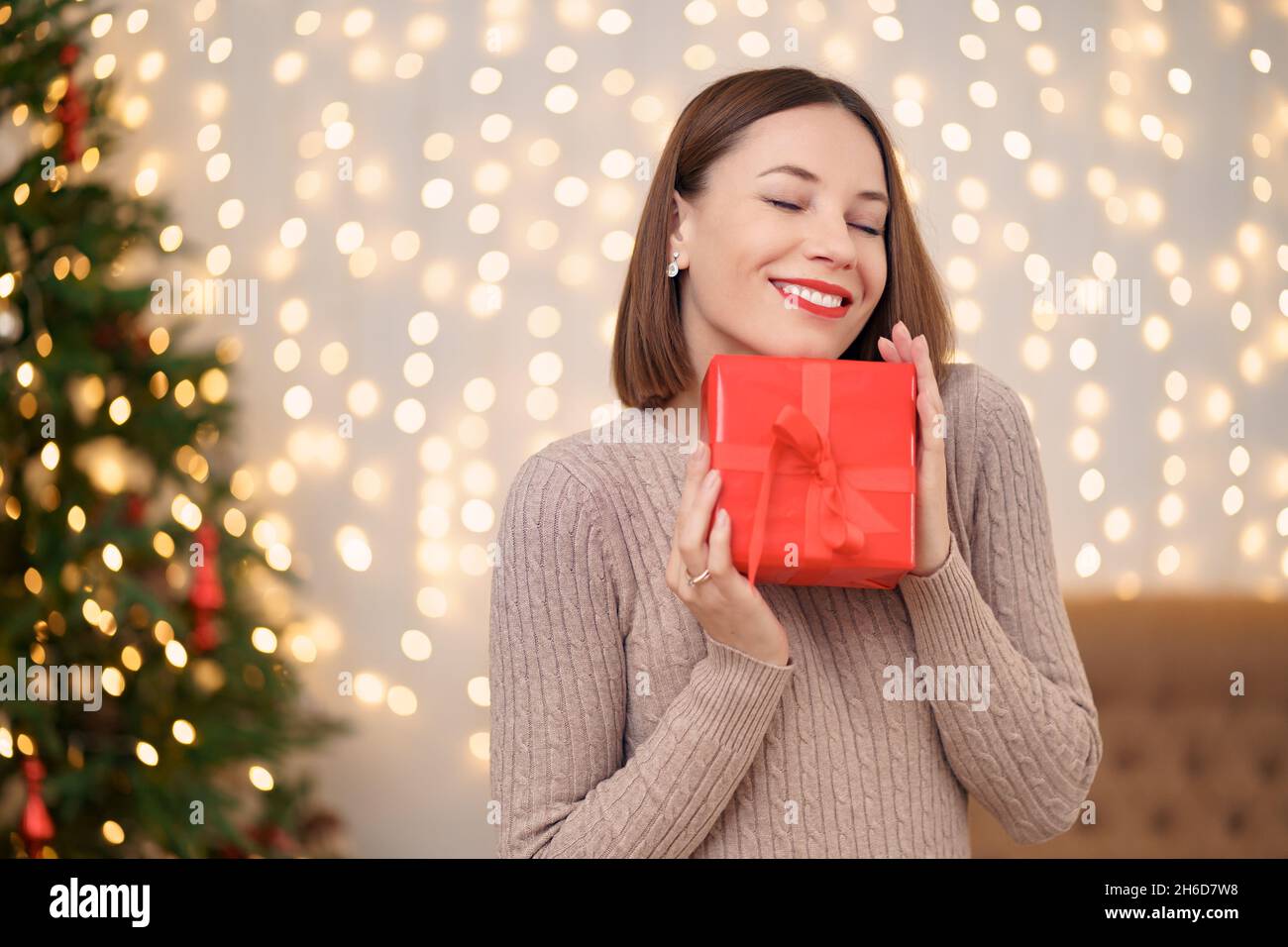 Portrait de la jeune femme heureuse lèvres rouges est si hapyy pour obtenir boîte cadeau enveloppée.Gros plan la femme satisfaite a reçu la boîte actuelle.Arrière-plan des lumières de Noël festives. Banque D'Images