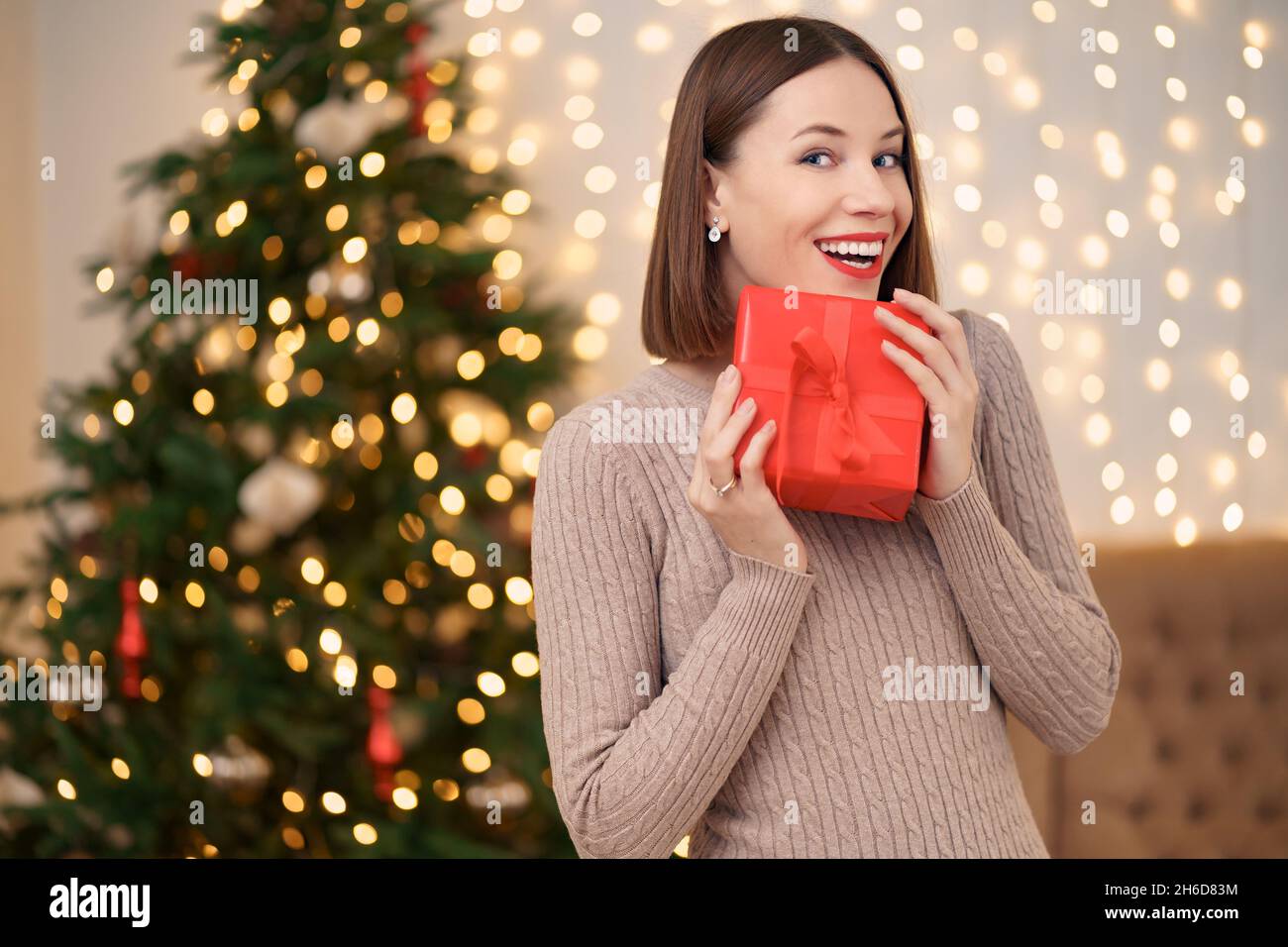 Portrait de jeune femme heureuse lèvres rouges posant avec une boîte cadeau emballée.Gros plan la femme satisfaite a reçu la boîte actuelle.Arrière-plan des lumières de Noël festives. Banque D'Images
