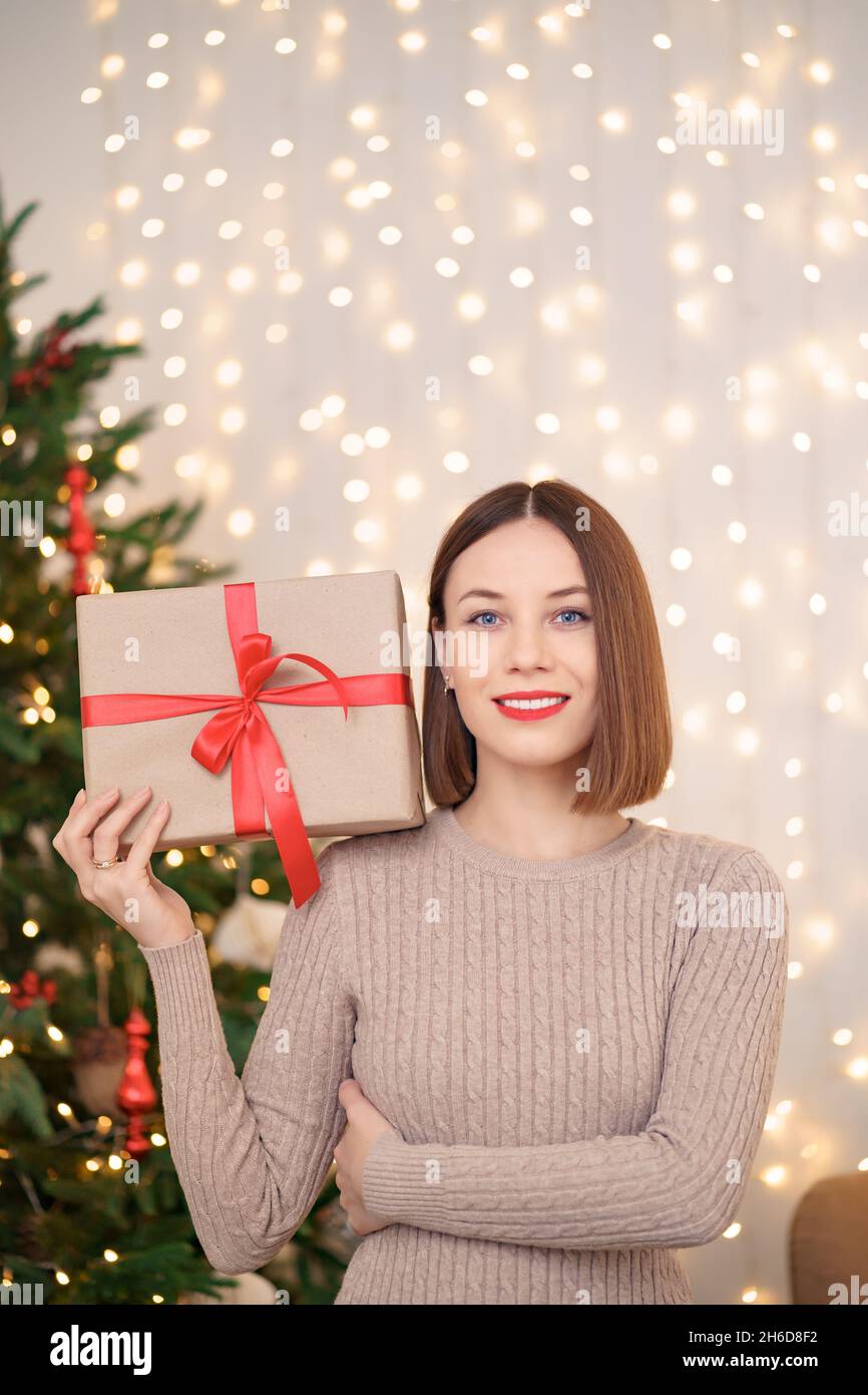 Portrait de jeune femme heureuse lèvres rouges regardant l'appareil photo tenant une boîte cadeau emballée.Gros plan la femme satisfaite a reçu la boîte actuelle.Arrière-plan des lumières de Noël festives. Banque D'Images