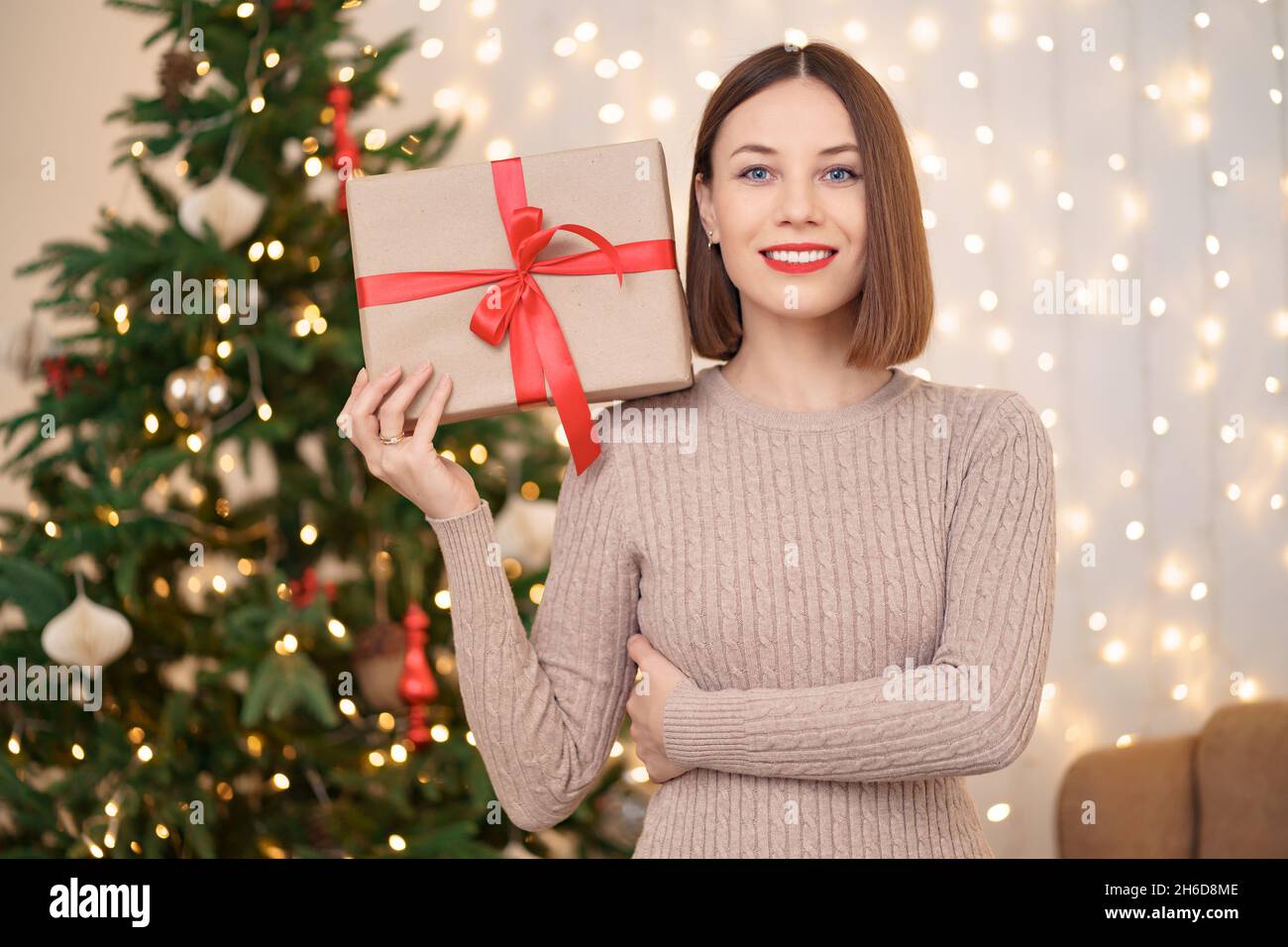 Portrait de jeune femme heureuse lèvres rouges regardant l'appareil photo tenant une boîte cadeau emballée.Gros plan la femme satisfaite a reçu la boîte actuelle.Arrière-plan des lumières de Noël festives. Banque D'Images