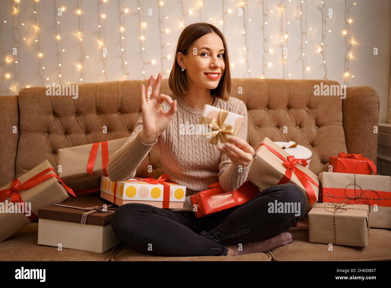 Bonne jeune femme tenant un cadeau montrant le signe ok entouré de boîtes-cadeaux assis jambes croisées sur un canapé de chameau avec des lumières en arrière-plan Banque D'Images