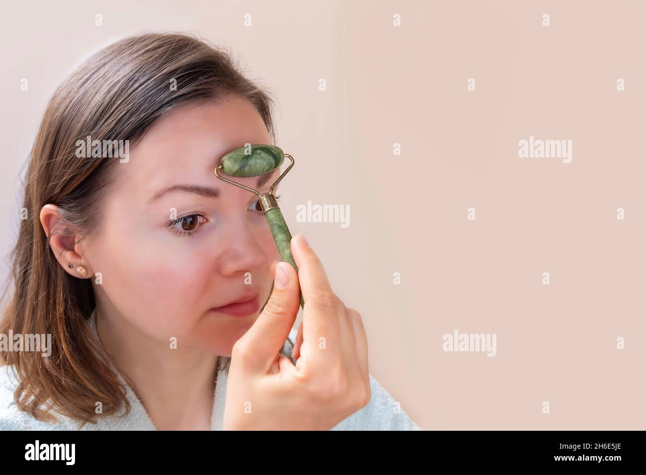 Jeune femme caucasienne effectuant un massage du visage avec un rouleau de jade vert sur fond neutre Banque D'Images