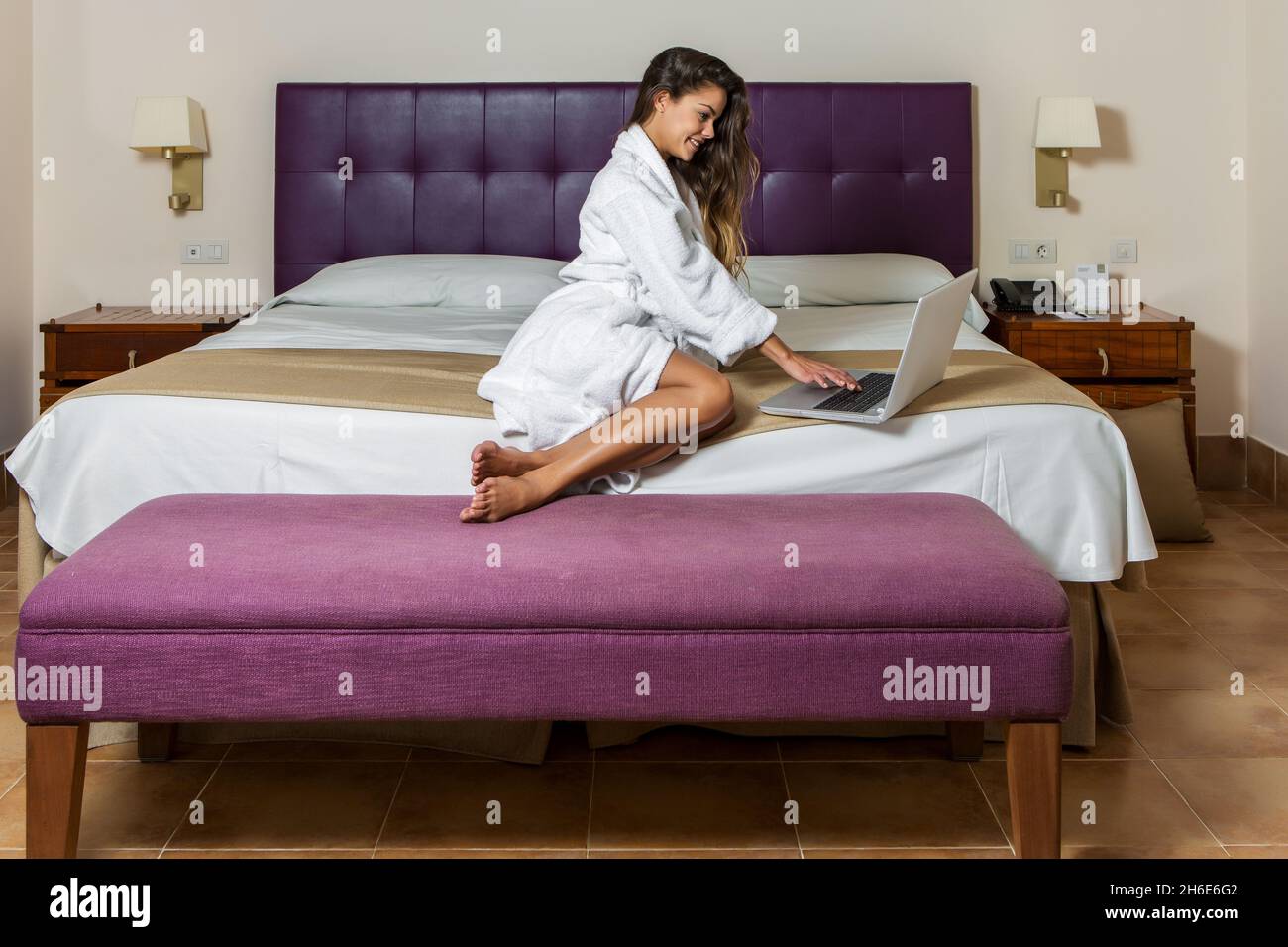 Femme souriante dans l'hôtel peignoir assis sur le lit dans un intérieur élégant et utilisant l'ordinateur portable heureusement Banque D'Images