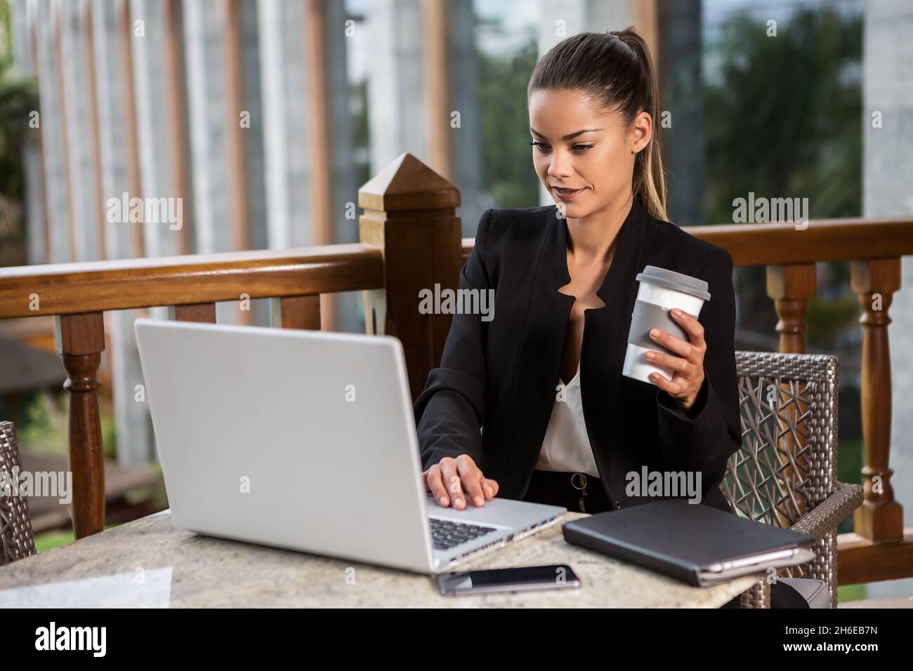 Jolie femme moderne dans un élégant costume de surf ordinateur portable et de prendre une tasse de café tout en étant assis à une table sur la terrasse Banque D'Images
