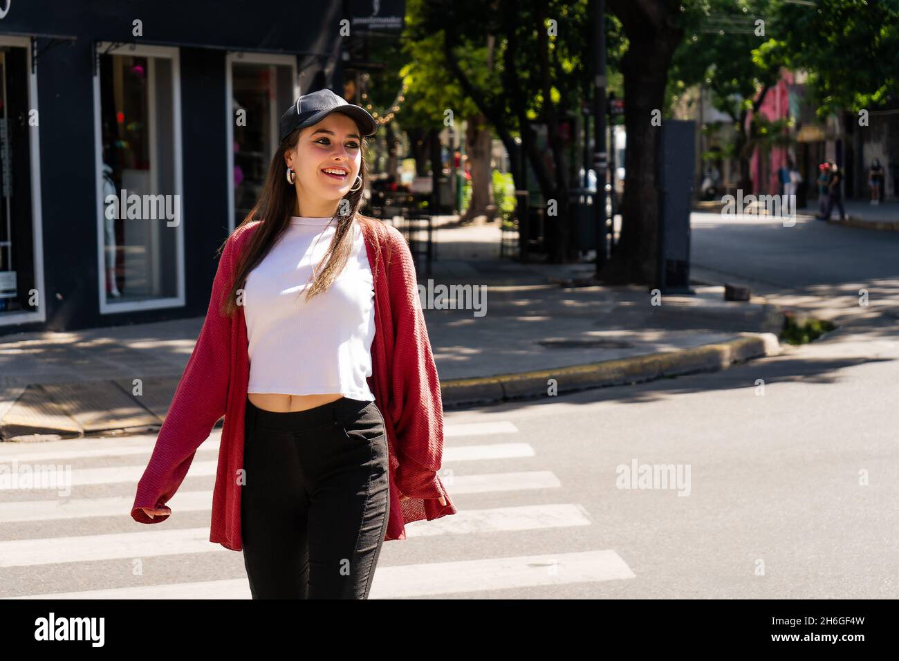 Une belle fille de Latina dans un chapeau noir traversant une rue sur le chemin piétonnier tout en souriant heureux. Banque D'Images