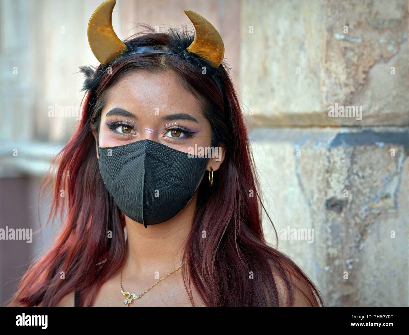 Jolie fille mexicaine avec des cornes de diable jaune serre-tête porte un masque noir pendant la pandémie mondiale de coronavirus et regarde le spectateur. Banque D'Images