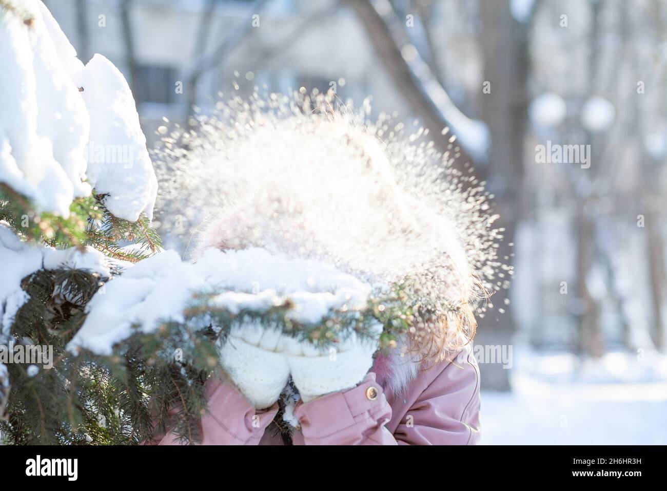 Une jeune femme en gants tricotés blancs souffle de la neige sur une branche, formant un nuage de neige.Arrière-plan hivernal avec bokeh et mise au point sélective Banque D'Images