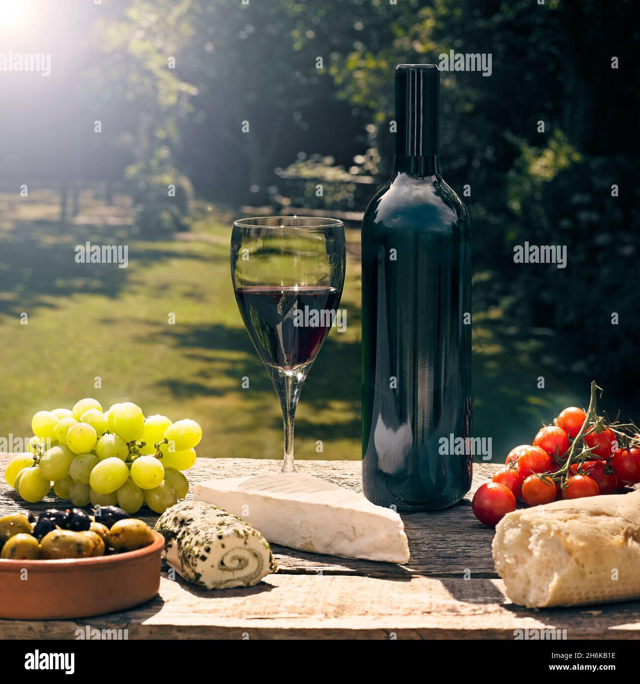 Bouteille de vin rouge, sans étiquette et verre plein de vin à table au soleil avec nourriture, fromages, olives, raisins et pain Banque D'Images