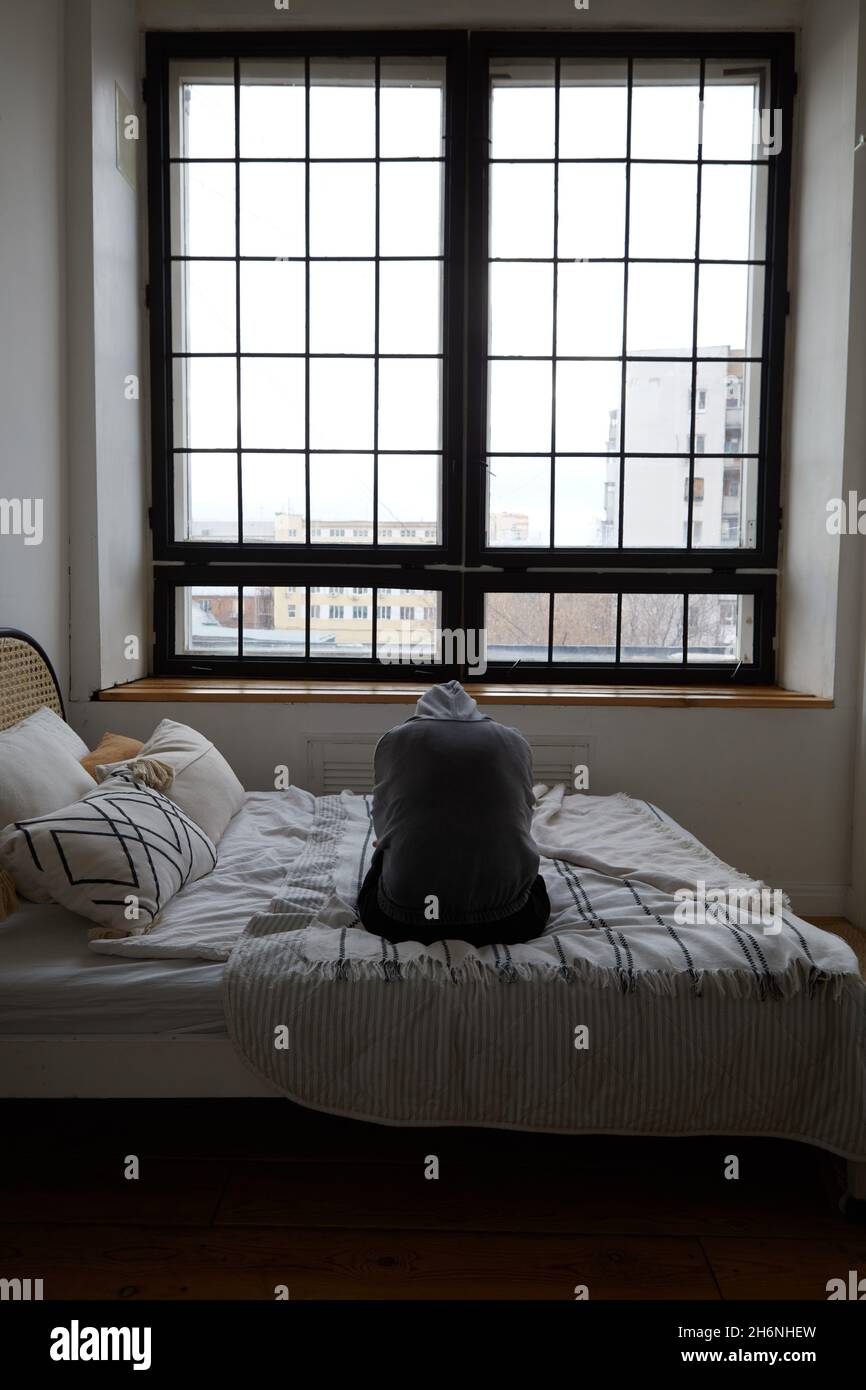 Vue arrière de la personne mélancolique anonyme dans la hotte souffrant d'anxiété assise sur le lit près de la fenêtre avec treillis Banque D'Images
