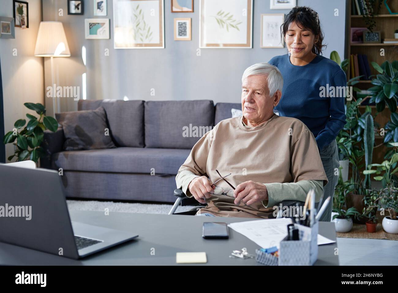 Femme mature qui s'occupe de l'homme âgé à la maison, elle le porte sur un fauteuil roulant jusqu'à la table avec un ordinateur Banque D'Images