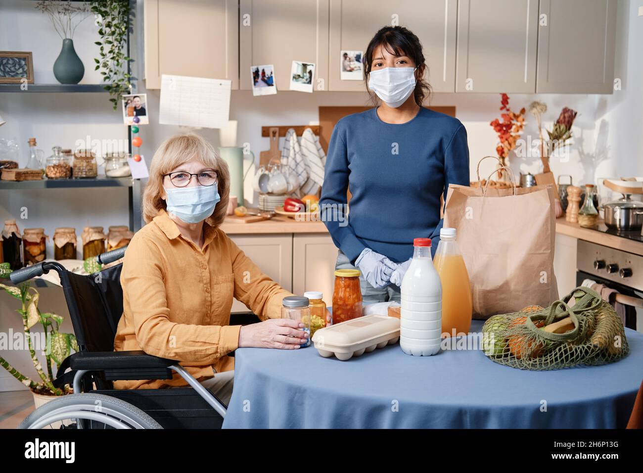 Portrait d'une femme âgée en fauteuil roulant assise à la table avec des produits et regardant l'appareil photo avec le soignant debout à proximité, ils portent des masques Banque D'Images