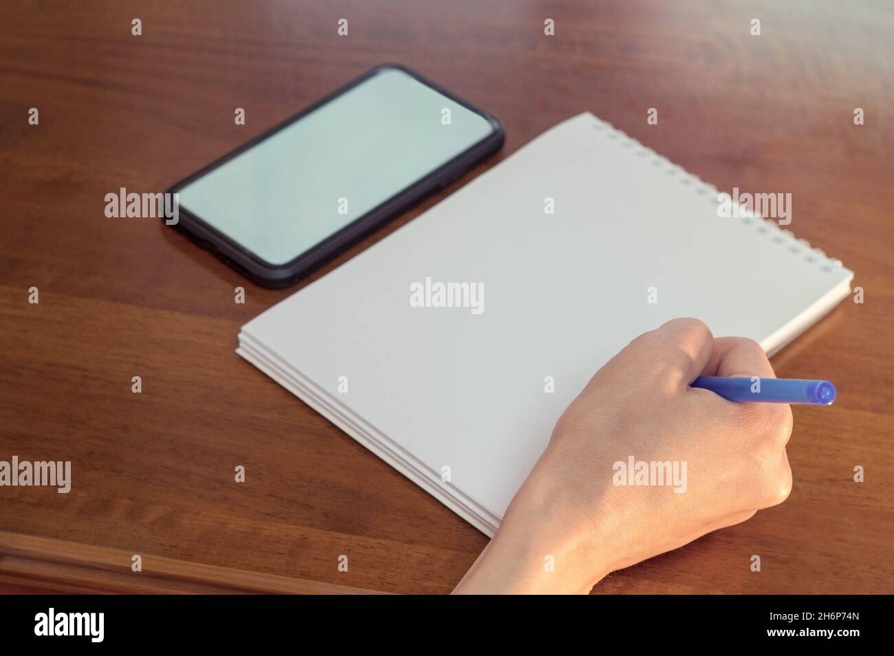 Une femme écrit dans un bloc-notes.Un téléphone cellulaire avec un écran vierge sur l'arrière-plan d'une table en bois.Espace de travail avec bloc-notes et stylet.espace de copie de maquette. Banque D'Images