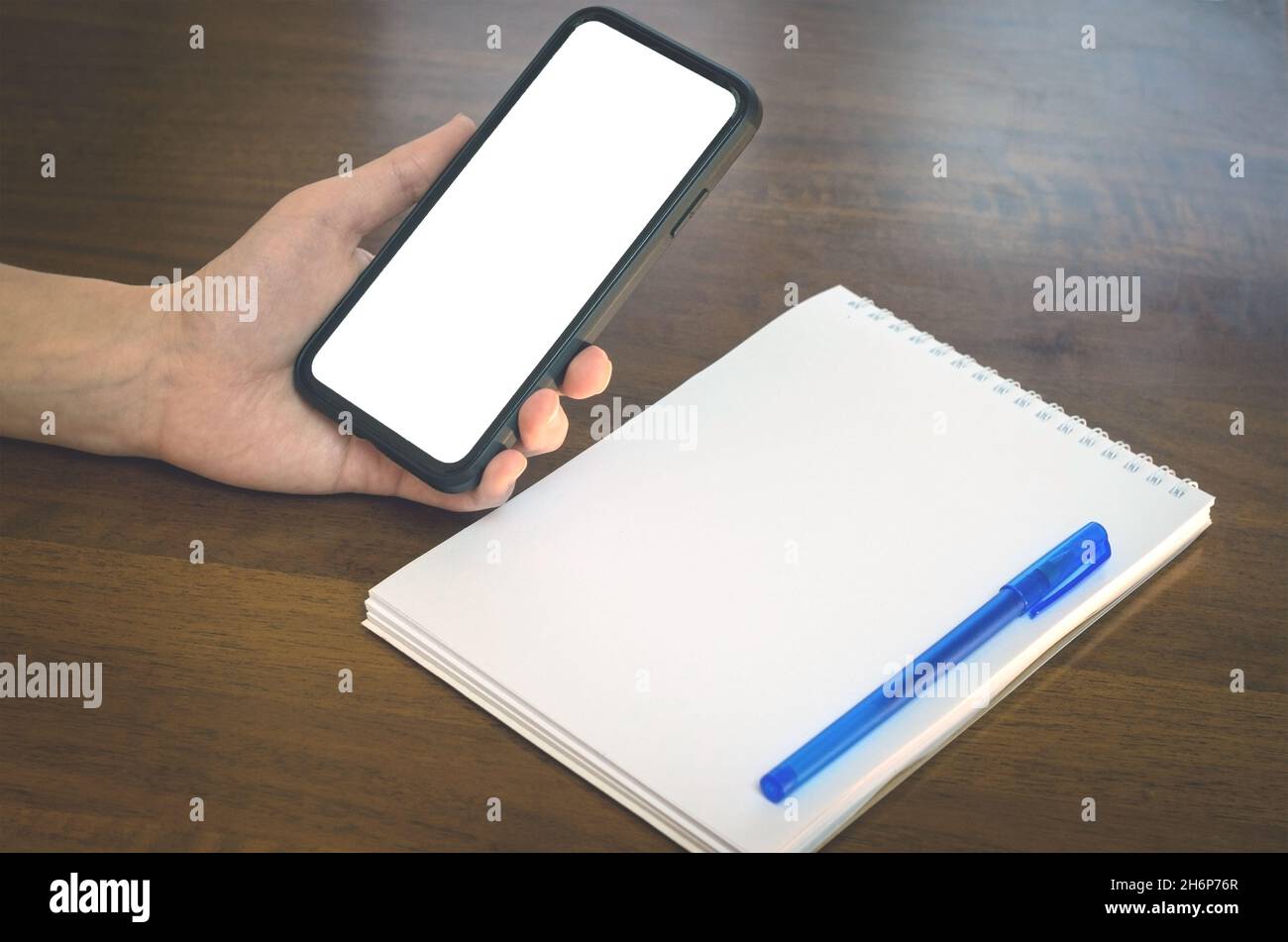 Une femme tient un téléphone cellulaire avec un écran vide sur le fond d'une table en bois.Espace de travail avec bloc-notes et stylet.espace de copie de maquette. Banque D'Images