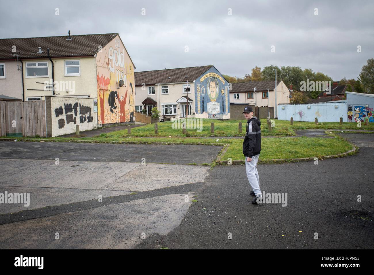 Jeune homme marchant devant des peintures murales politiques avec des symboles paramilitaires sur le Lower Shankill Estate, Belfast. Banque D'Images