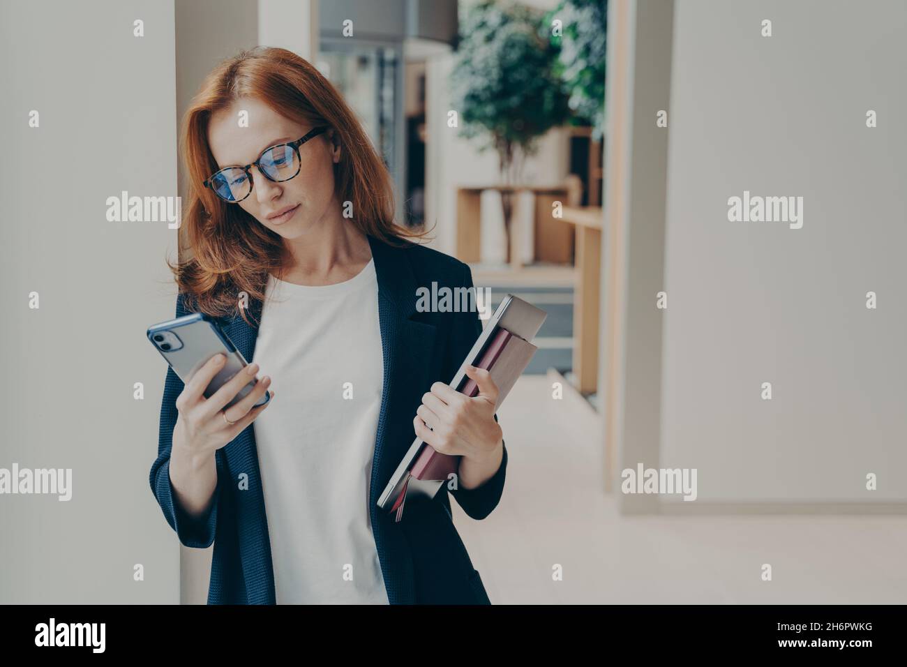 REDHEAD femme d'affaires avec un regard sérieux dans les lunettes en utilisant le téléphone cellulaire en se tenant au bureau Banque D'Images