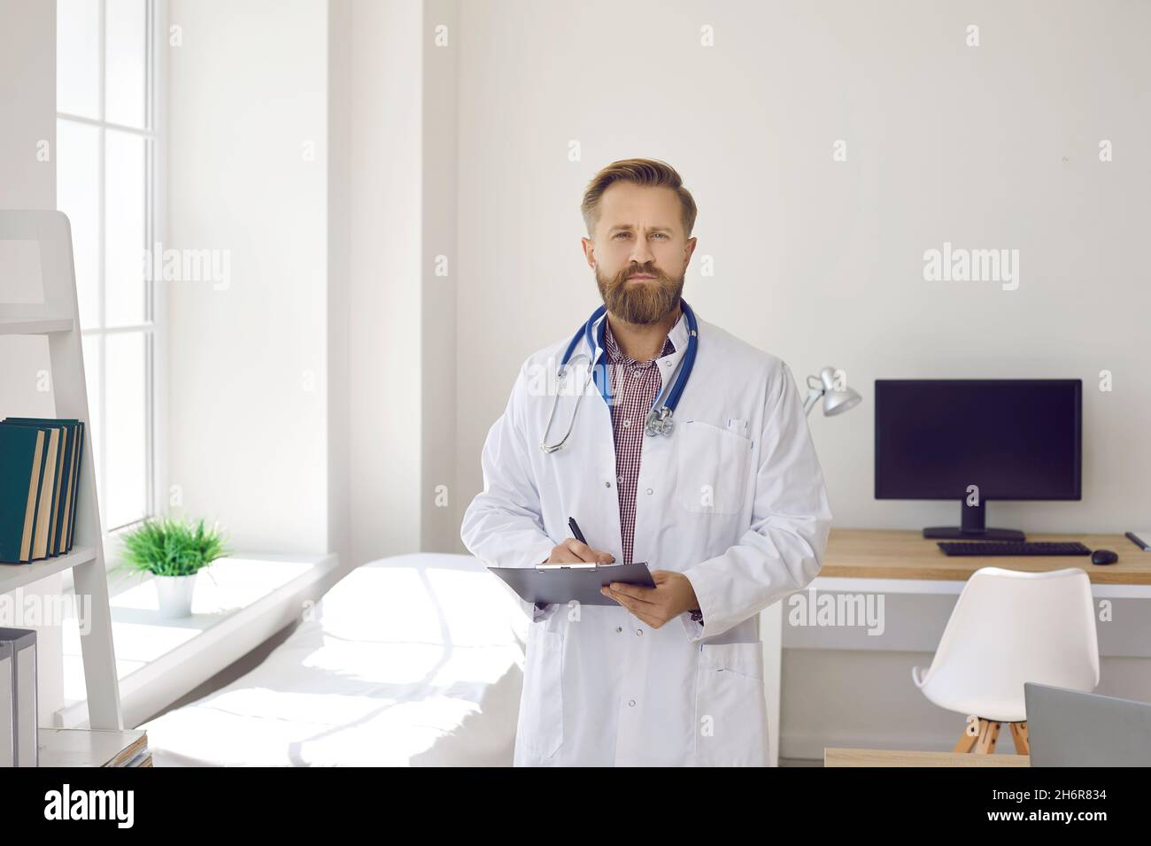 Portrait d'un médecin masculin sérieux debout dans un bureau médical et tenant une planchette à pince Banque D'Images