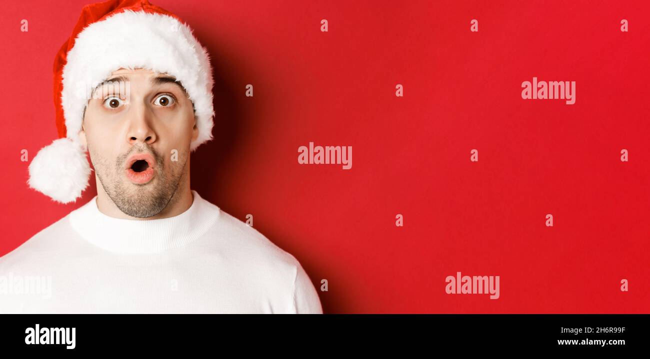 Gros plan d'un joli homme surpris dans le chapeau de santa, en disant wow  et regardant quelque chose d'intéressant, debout sur fond rouge Photo Stock  - Alamy