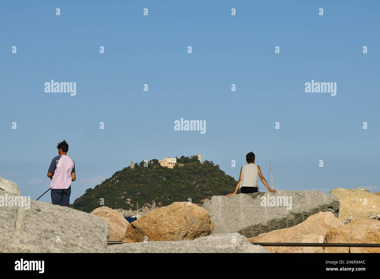 Deux garçons de derrière pêchent et se détendent sur les rochers au bord de la mer avec l'île de Gallinara en arrière-plan en été, Alassio, Savona, Ligurie Banque D'Images
