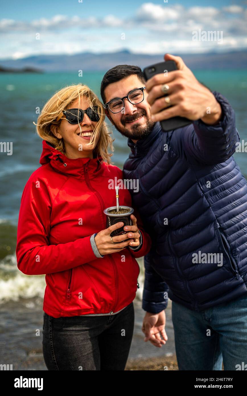 Petit foyer d'un couple argentin souriant prenant un selfie à la plage par une journée venteuse Banque D'Images