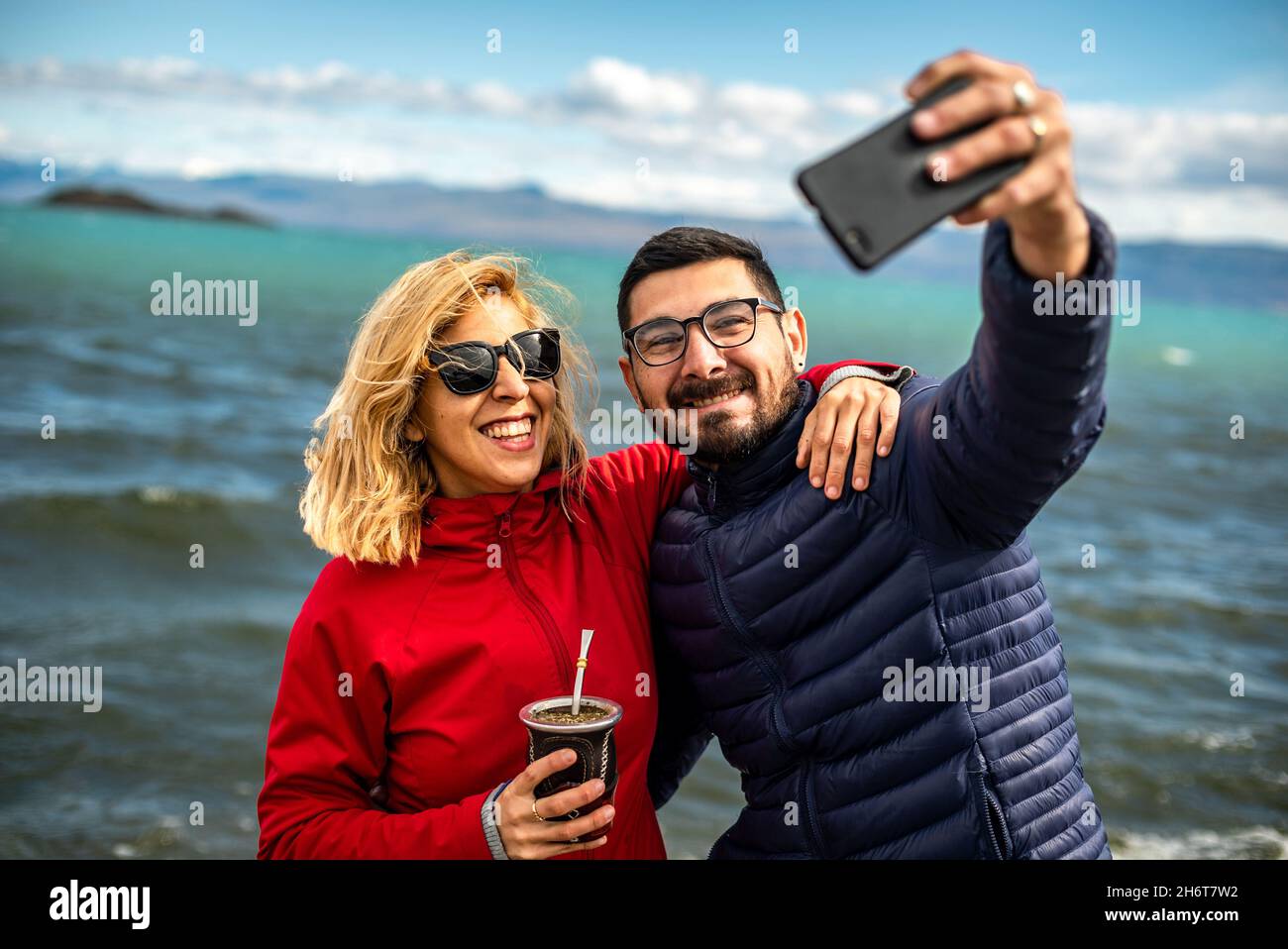 Petit foyer d'un couple argentin souriant prenant un selfie à la plage par une journée venteuse Banque D'Images