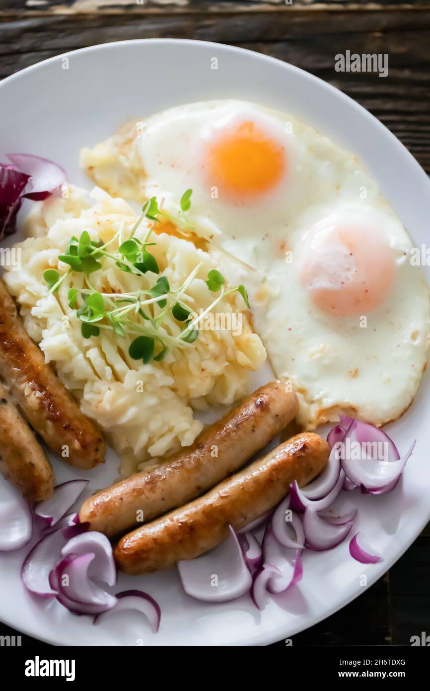 Saucisses de porc avec oignons, œufs, purée de pommes de terre et microlégumes.Petit déjeuner copieux.Fond en bois foncé, plaque en céramique blanche.Vue de dessus. Banque D'Images