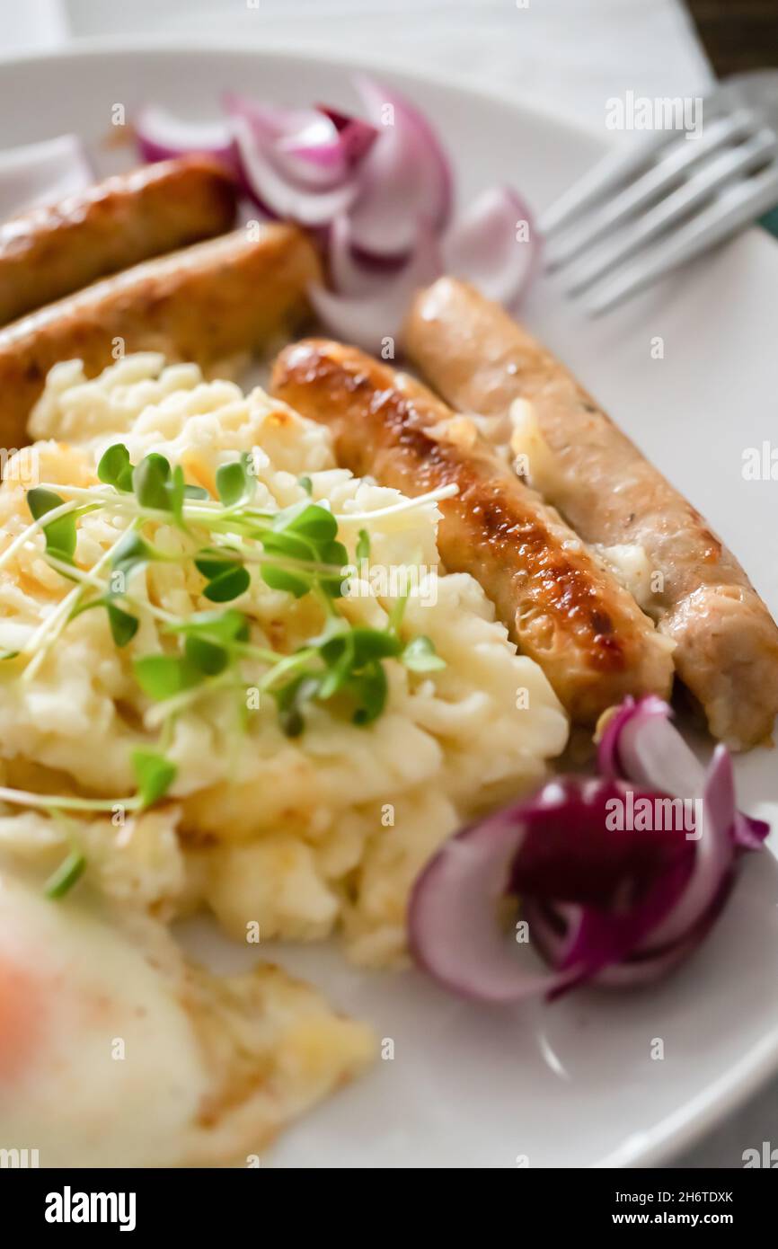 Saucisses de porc avec oignons, œufs, purée de pommes de terre et microlégumes.Petit déjeuner copieux.Fond en bois foncé, plaque en céramique blanche.Vue de dessus. Banque D'Images