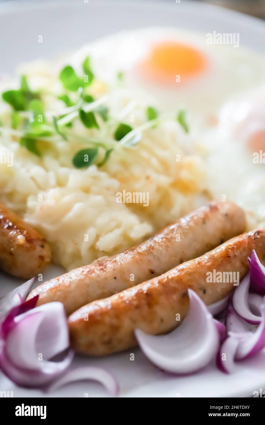 Saucisses de porc avec oignons, œufs, purée de pommes de terre et microlégumes.Petit déjeuner copieux.Fond en bois foncé, plaque en céramique blanche.Vue de dessus. Banque D'Images