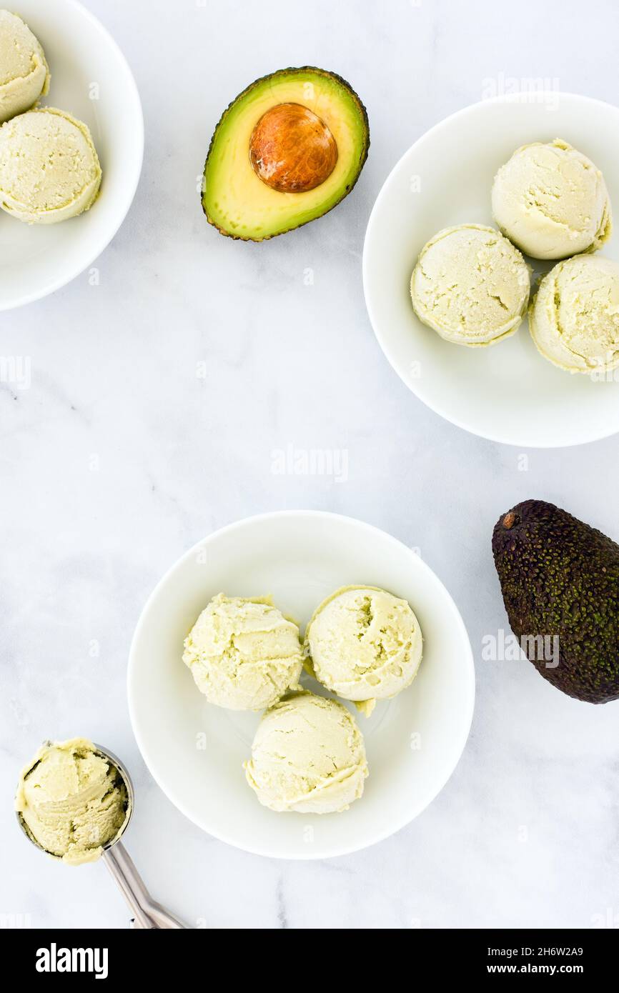 Crème glacée à l'avocat dans trois bols, cuillères, cuillère à crème glacée et moitié d'avocat sur marbre blanc avec espace de copie.Vue de dessus. Banque D'Images