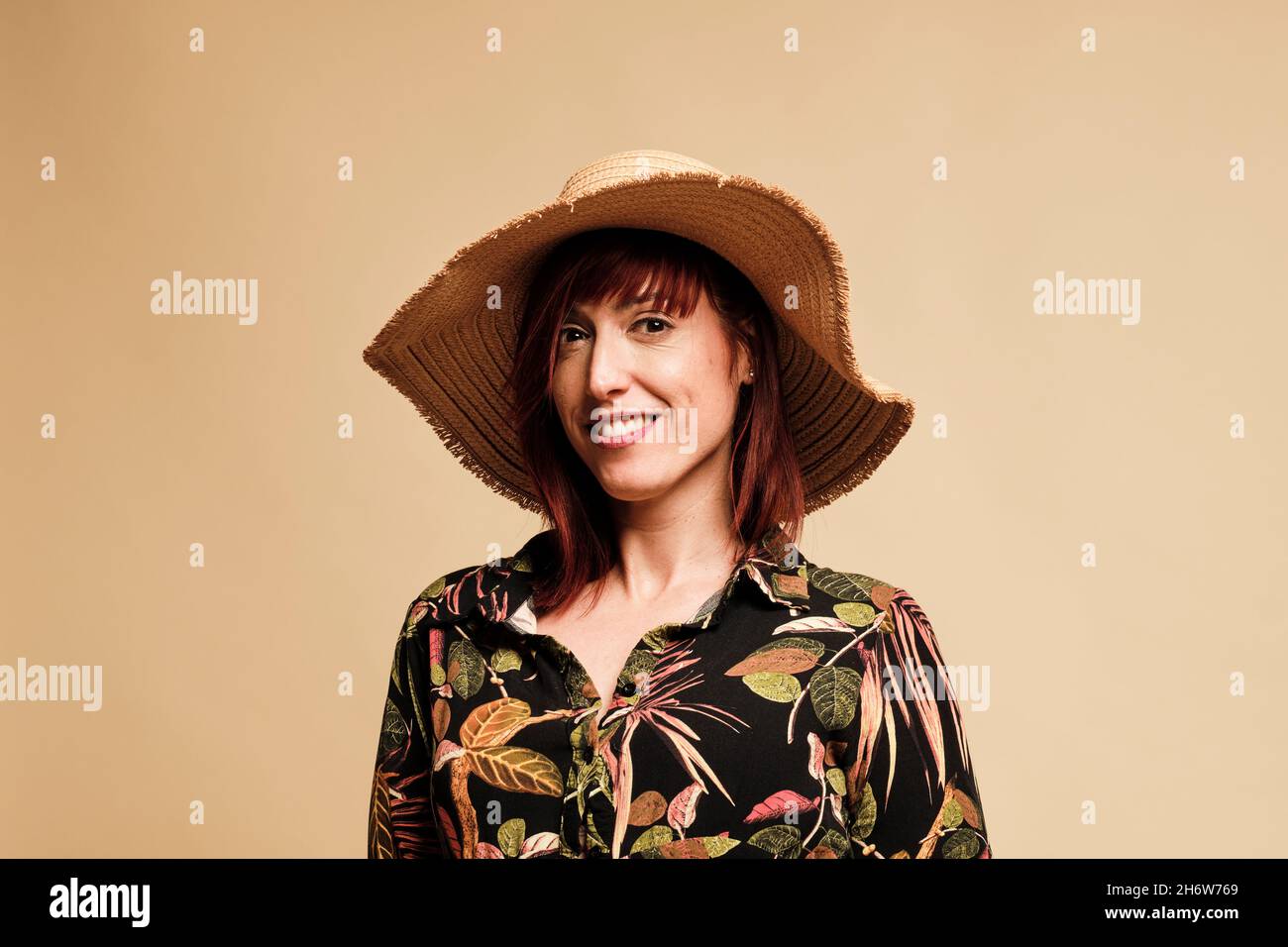 Jolie femme à tête rouge souriant avec une longue robe à fleurs et un chapeau.Portrait, avec un arrière-plan jaune. Banque D'Images