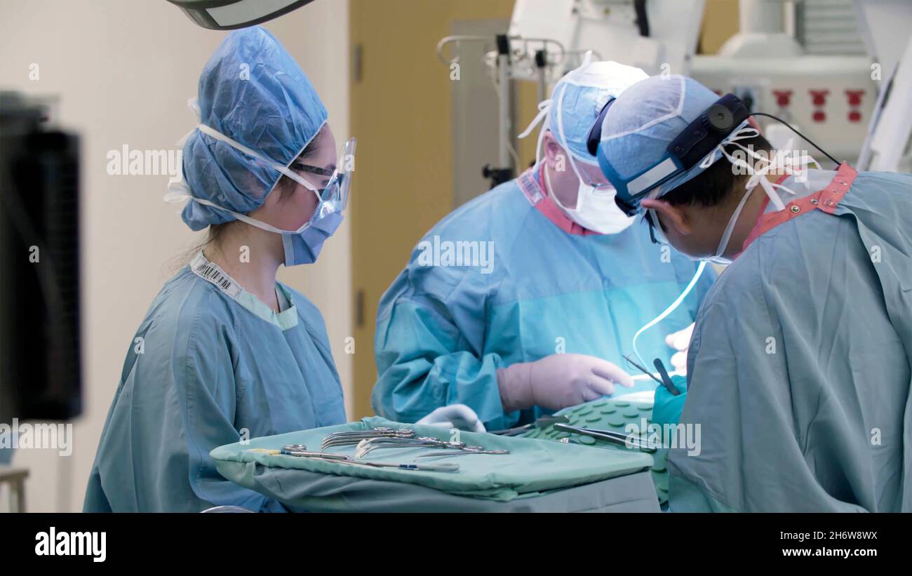 gros plan des médecins travaillant sur un patient dans une salle d'opération avec un étudiant ou une infirmière observant, et une table d'outils chirurgicaux à côté. Banque D'Images