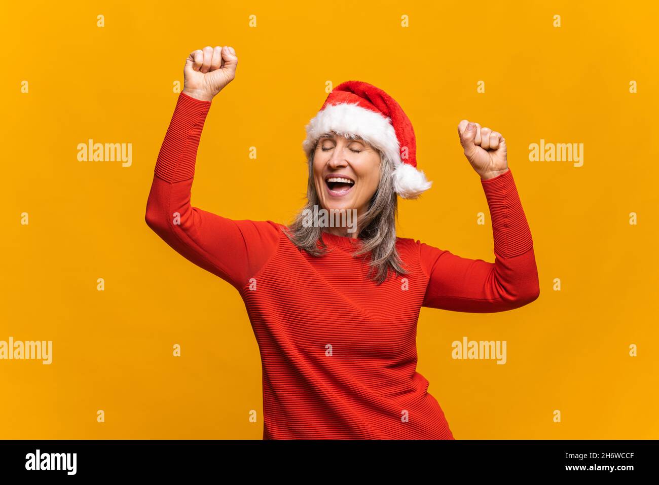 Folle femme moderne mature portant chapeau de père noël dansant avec les yeux fermés dans ecstatic isolé sur fond jaune.Femme sénior à cheveux gris célébrant la fête du nouvel an, Noël.Concept publicitaire Banque D'Images