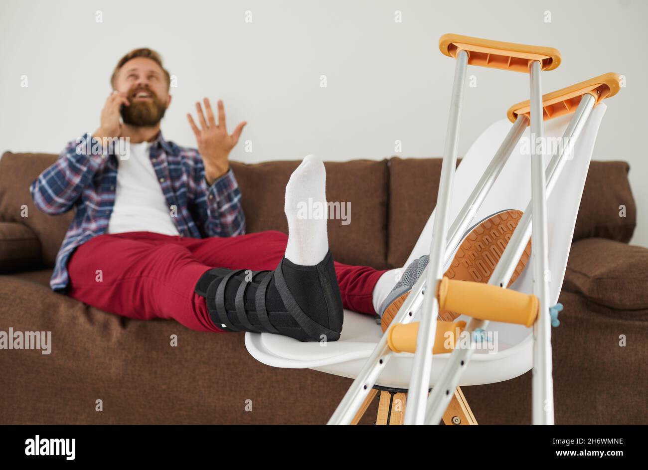 Homme avec une jambe cassée, un pied blessé ou une cheville tentaculaire assis sur un canapé et parlant sur un téléphone portable Banque D'Images