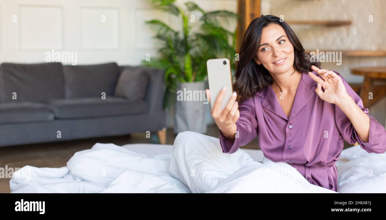 Photo d'une femme d'âge moyen au lit avec un téléphone portable.Elle se réveille faire selfie pour les publications de médias sociaux ou de blog.Technologie moderne et addict numérique Banque D'Images