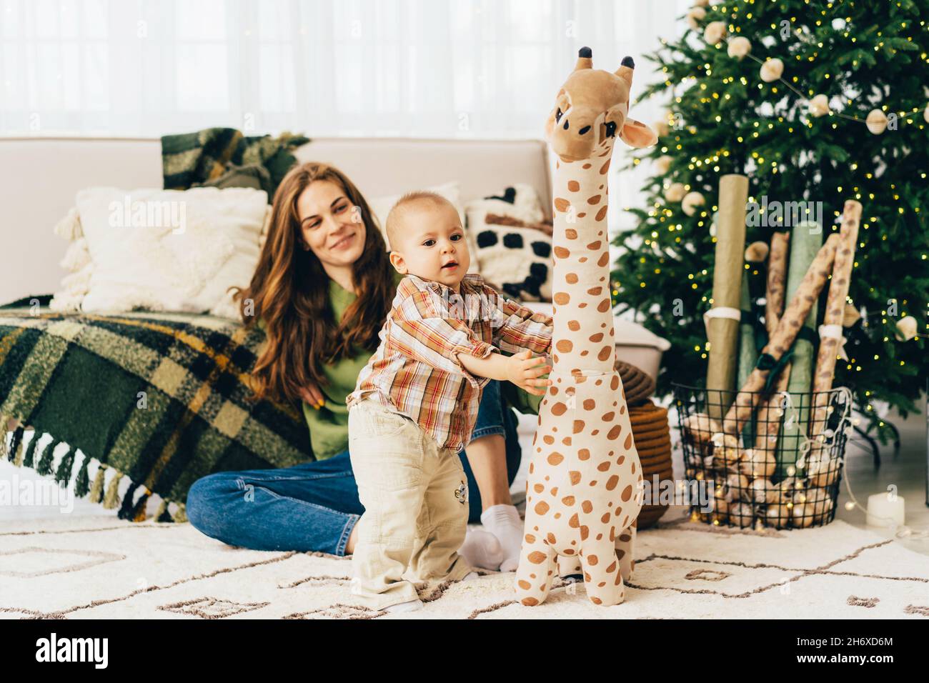 La momie aimante est assise sur le sol et regarde l'enfant jouer avec un grand jouet girafe en peluche. Banque D'Images