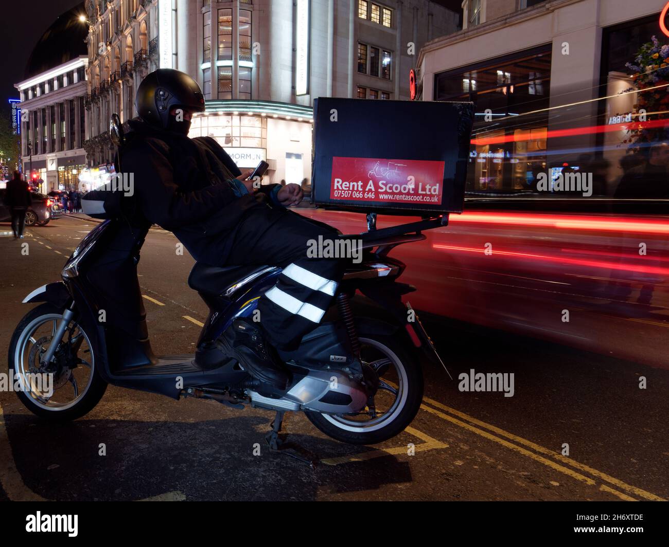 L'homme se penche sur son scooter en vérifiant son téléphone alors que les voitures passent en laissant des sentiers lumineux dans une longue exposition. Londres Banque D'Images