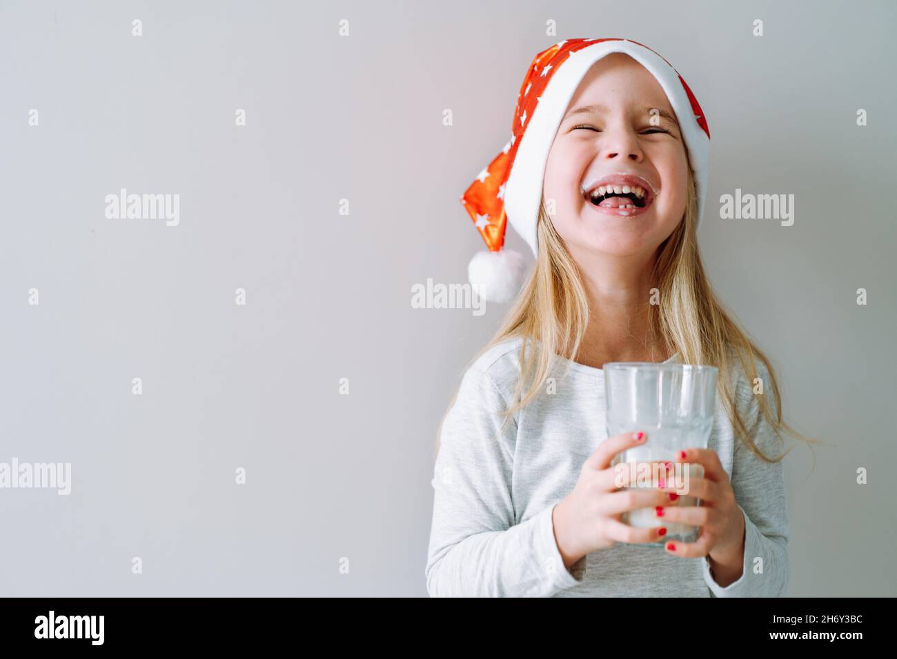 Portrait d'une petite fille en pyjama et chapeau de père noël riant avec verre de lait sur fond clair.Espace pour le texte Banque D'Images