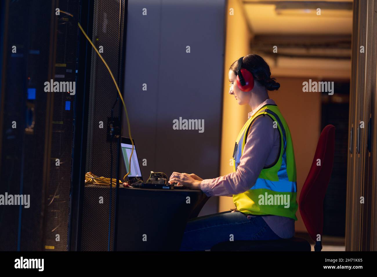 Femme d'ingénieur caucasien portant des bouchons d'oreille à l'aide d'un ordinateur portable dans la salle des serveurs informatiques Banque D'Images