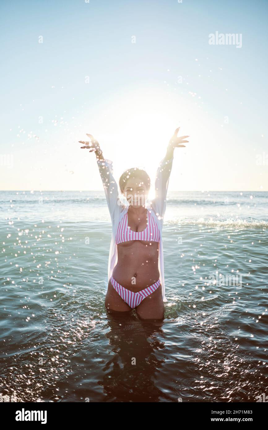 Sexy belle fille latine ayant un bain et jouant avec l'eau sur une plage déserte portant un bikini rose et des lunettes de soleil roses. Banque D'Images