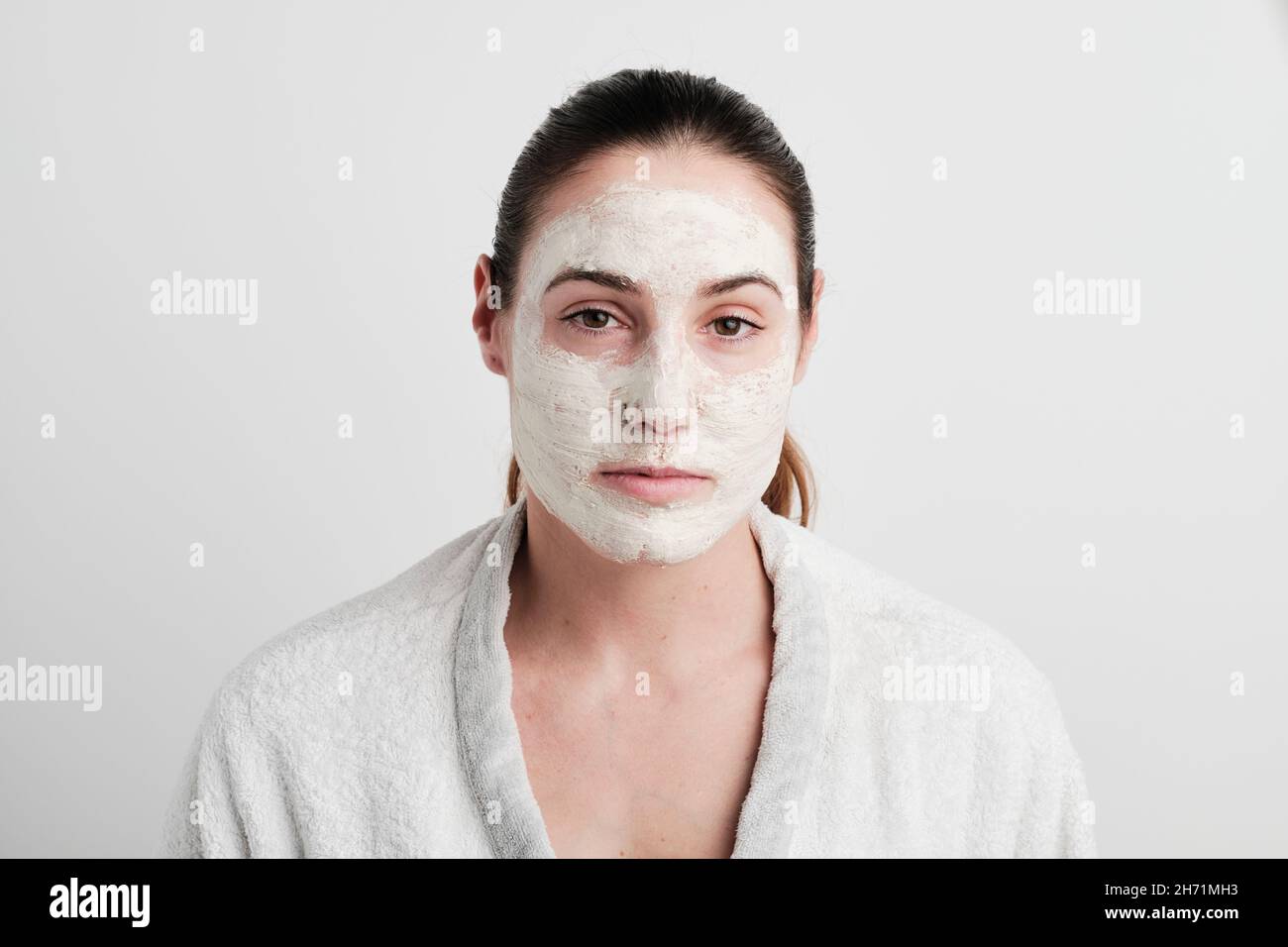 Jeune femme avec de la crème dans son visage regardant l'appareil photo portant un peignoir blanc sur un fond blanc Banque D'Images