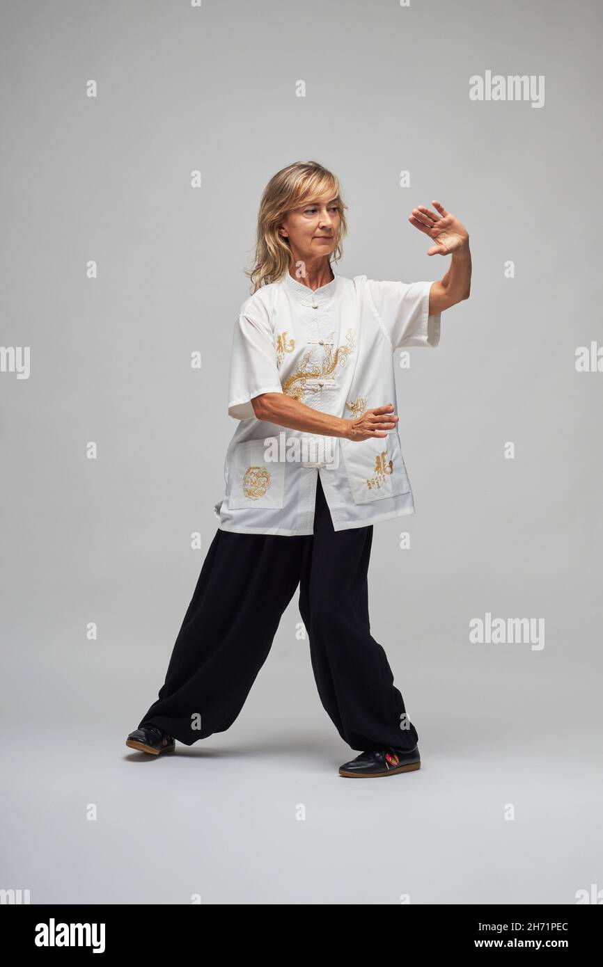 Femme blonde mature pratiquant le Chi kung et le Tai Chi sur fond blanc.Elle porte une veste chinoise traditionnelle de Tai Chi, un pantalon noir et de bl Banque D'Images