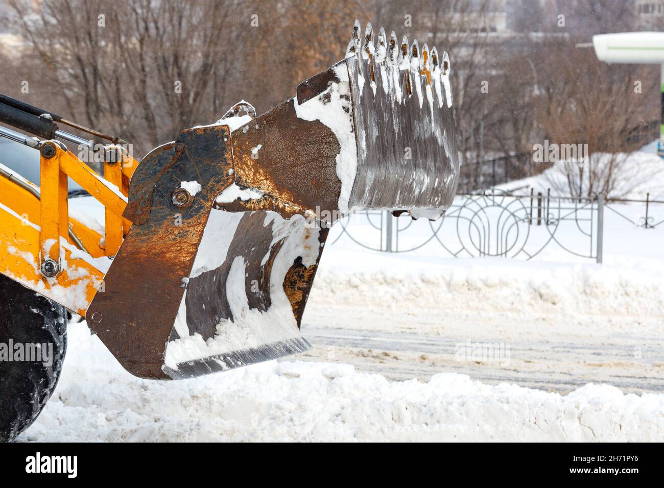 Un grand seau extensible d'un tracteur jaune pelle la neige d'une route urbaine sur fond de rue urbaine. Banque D'Images